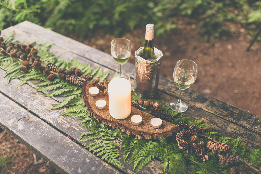 A bottle of wine and some glasses on a picnic bench decorated with ferns and pinecones